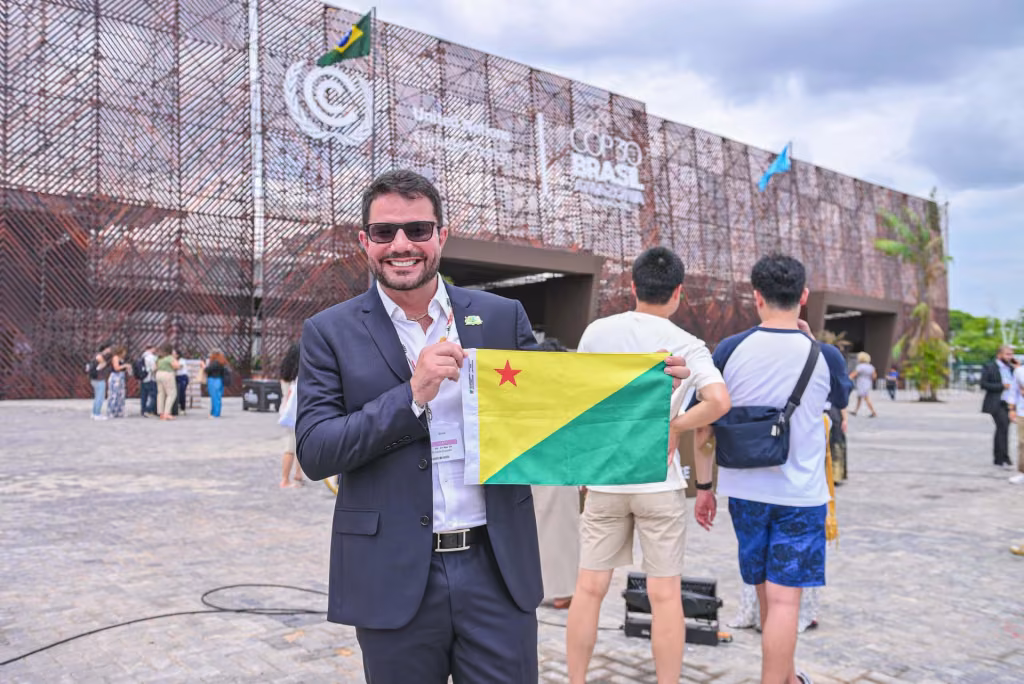 Gladson Cameli segura a bandeira do Acre em frente ao pavilhão de uma conferência internacional